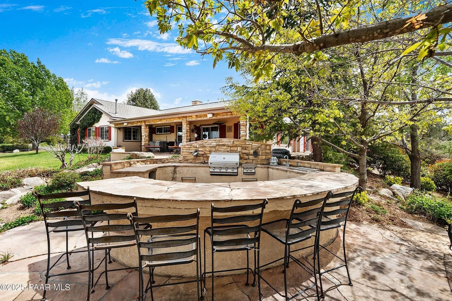 Outdoor patio with a circular stone bar, grill, and seating, surrounded by trees and greenery.