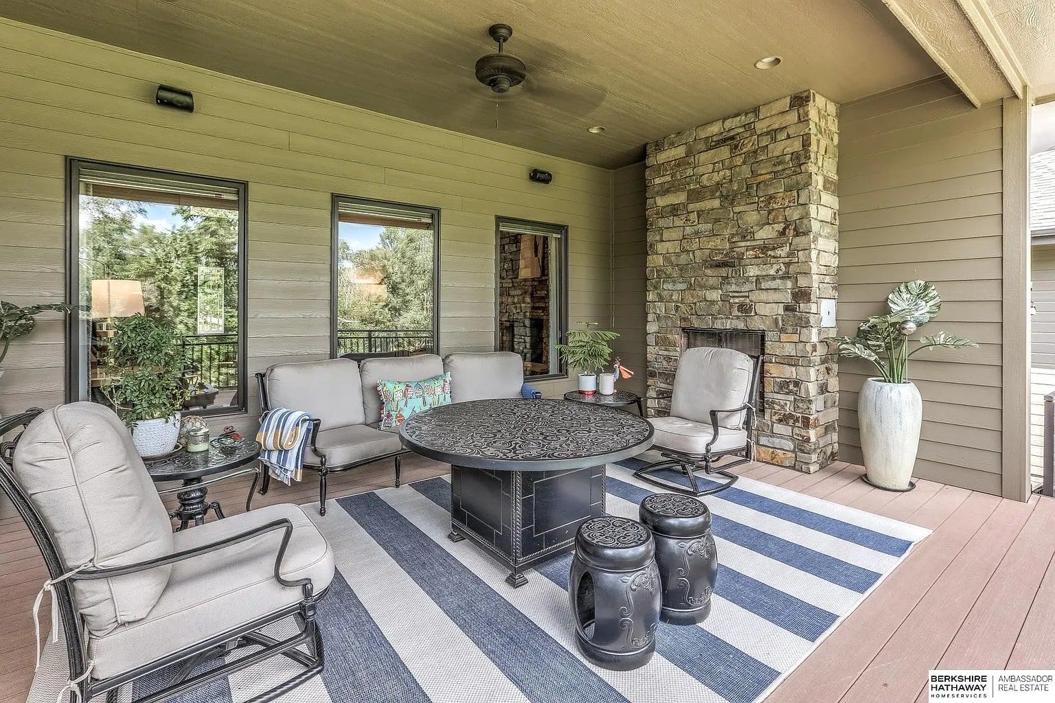Covered patio with cushioned chairs, striped rug, round table, and stone fireplace. Lush greenery outside.