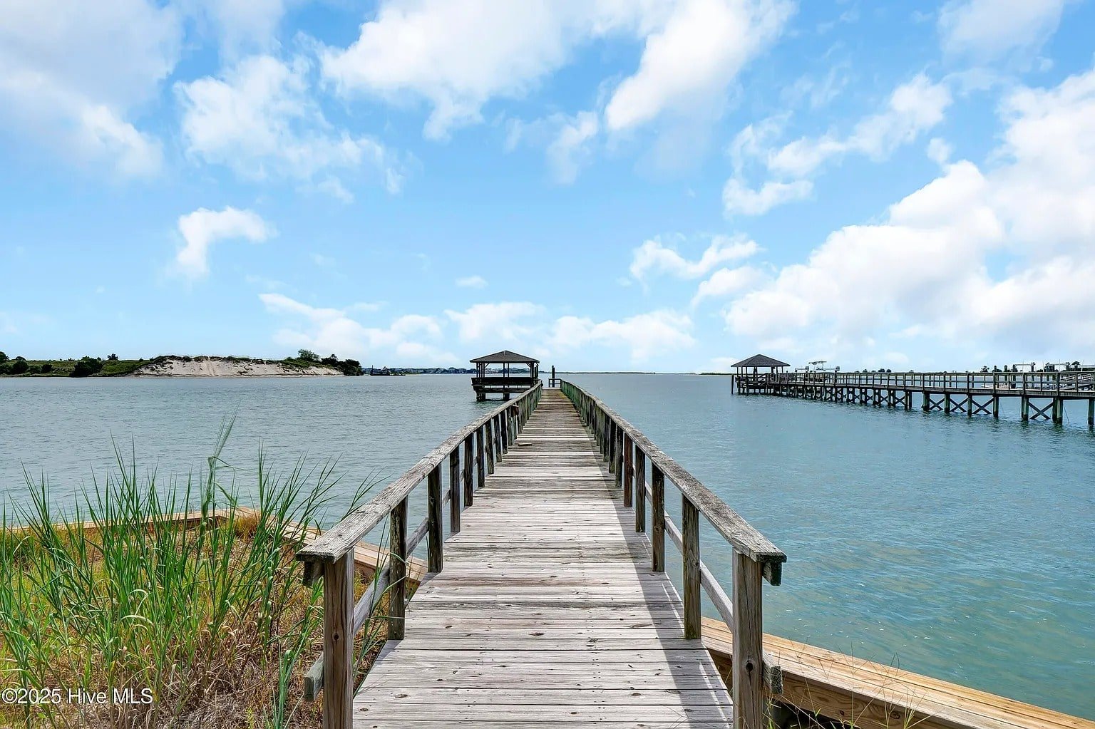 Wooden pier extending over calm water with two covered structures, under a partly cloudy sky.