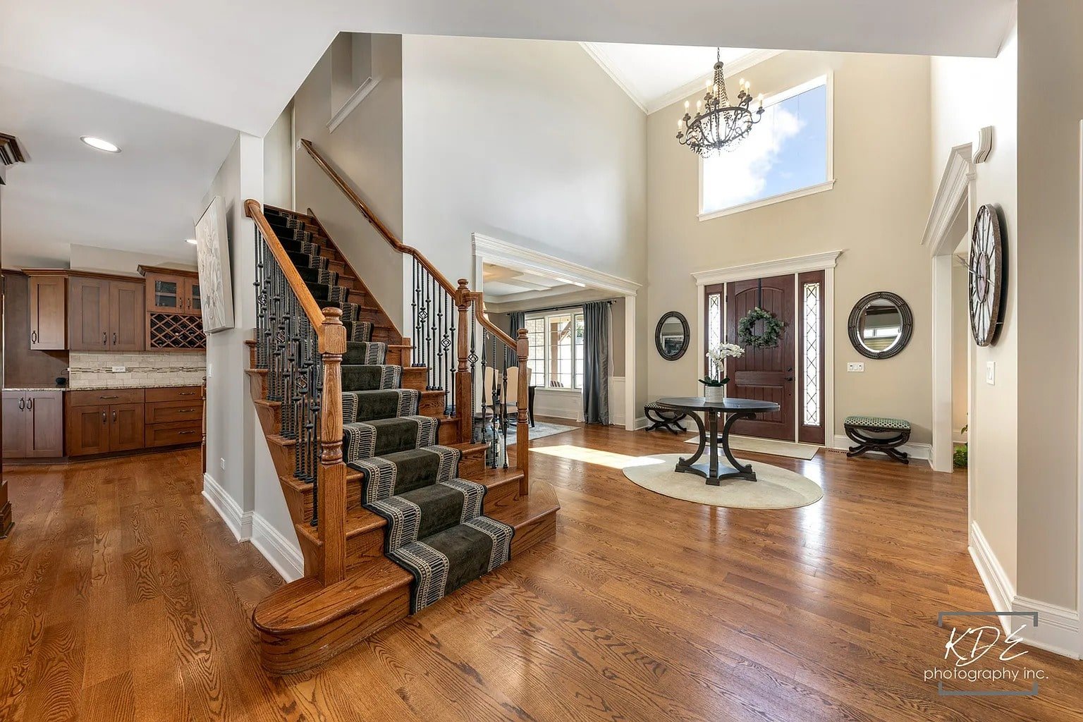 Spacious foyer with wooden staircase, chandelier, and round table, leading to kitchen and living area.