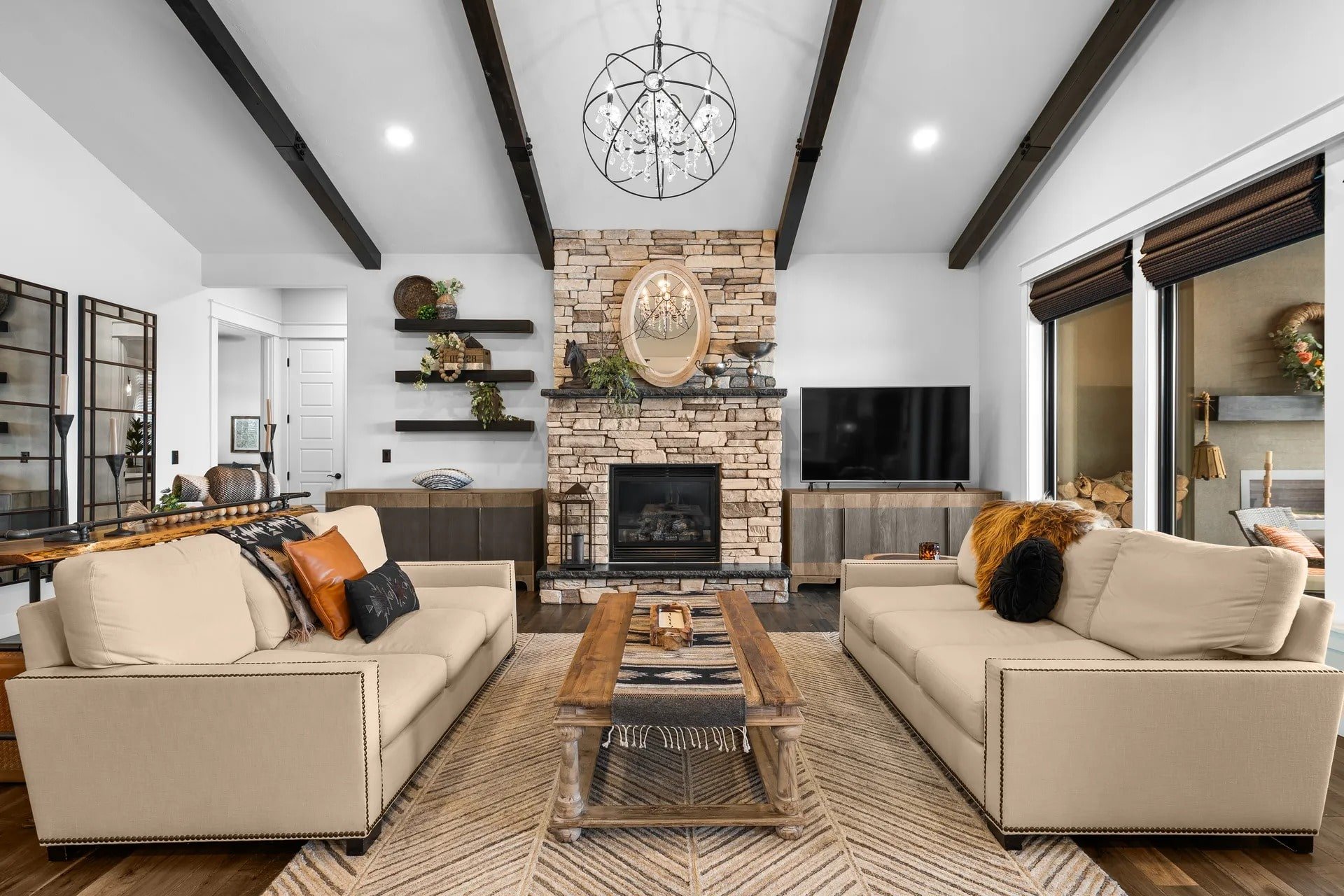 Modern living room with beige sofas, stone fireplace, wooden beams, and a large chandelier.