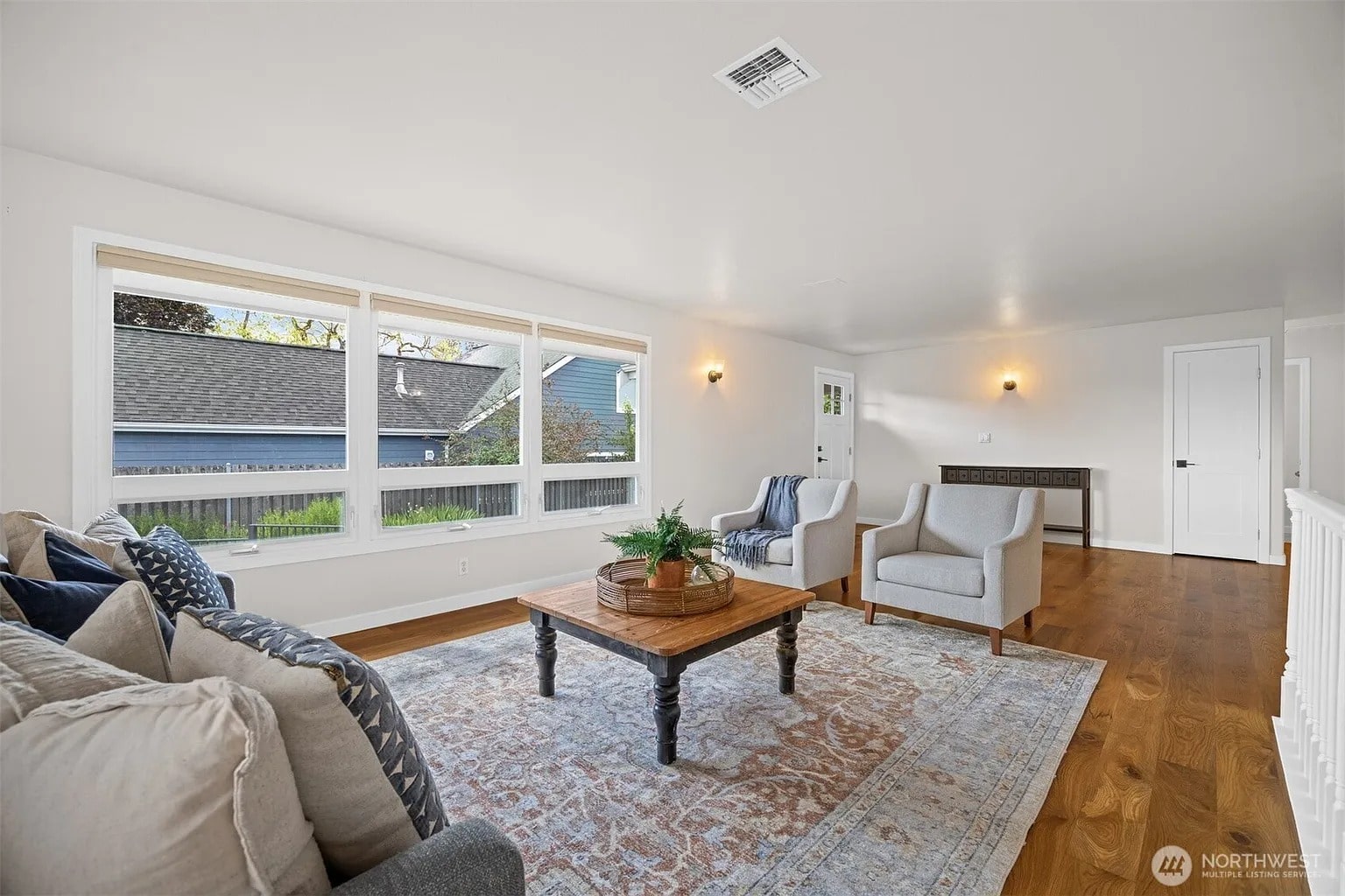 Bright living room with large windows, wooden floor, sofa, armchairs, and a decorative rug.