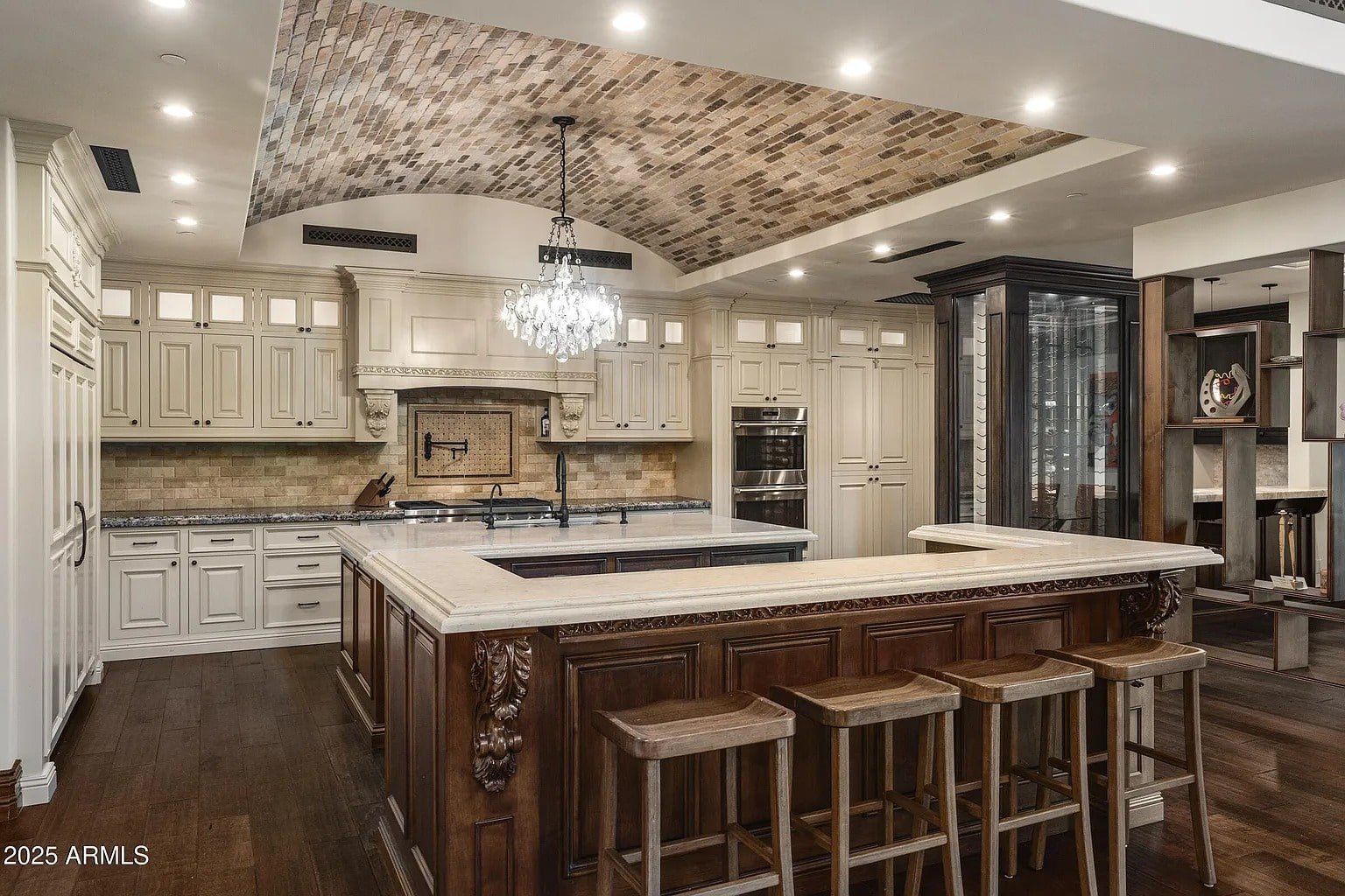 Luxurious kitchen with ornate wooden island, chandelier, and cream cabinetry under a brick-patterned ceiling.