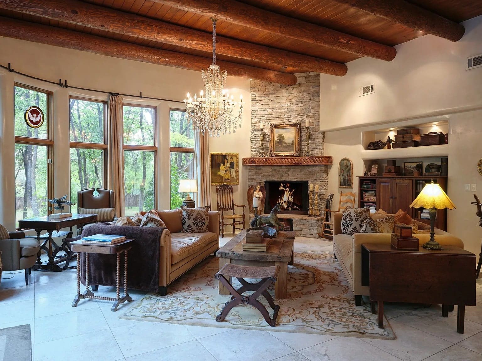 Rustic living room with wooden beams, stone fireplace, chandelier, and large windows overlooking greenery.