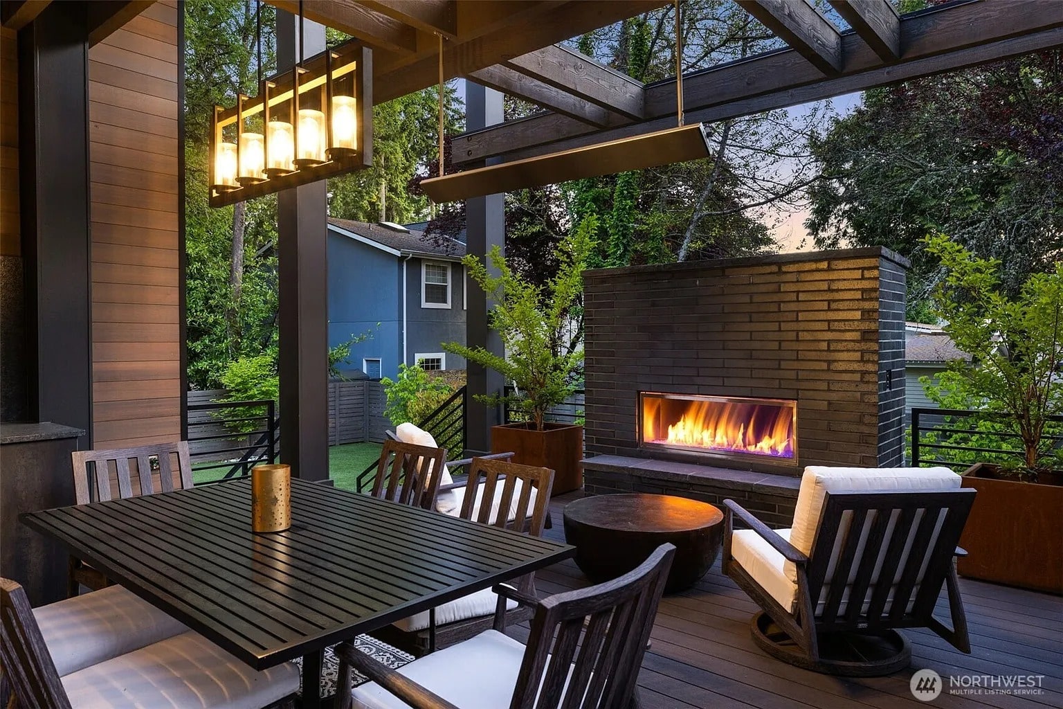Outdoor patio with dining table, chairs, and a lit fireplace under a pergola.