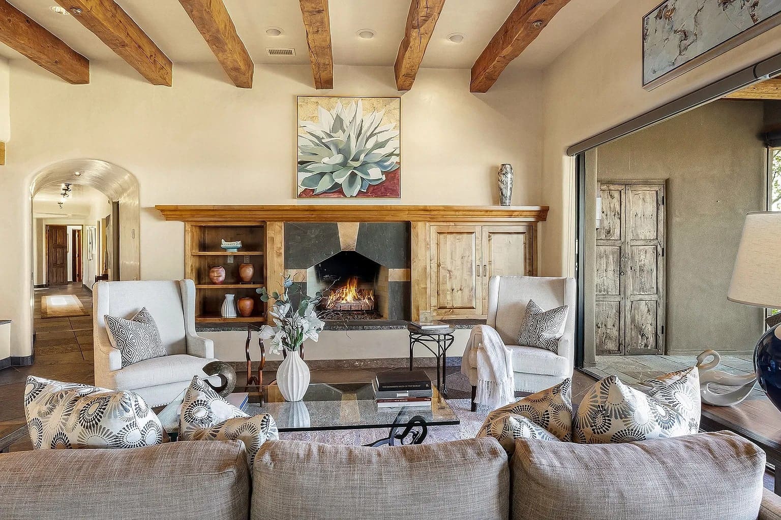 Cozy living room with a fireplace, wooden beams, and elegant furniture, featuring a large plant painting.