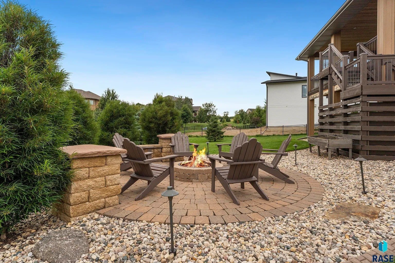 Outdoor patio with a circular fire pit, surrounded by wooden chairs and landscaped greenery.