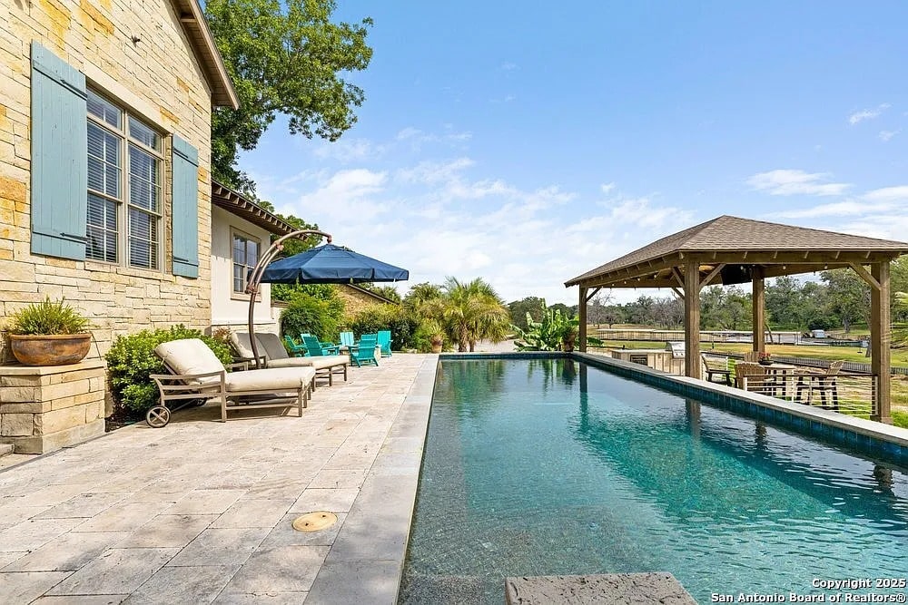 Luxury backyard with a stone patio, pool, lounge chairs, umbrella, and a wooden gazebo.