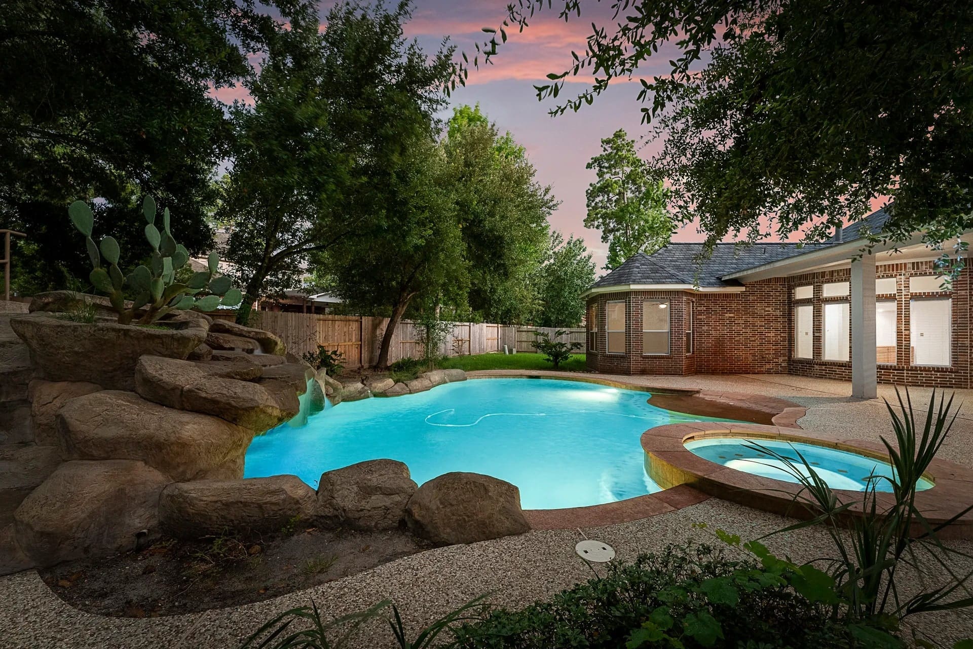 Backyard with a lit swimming pool, hot tub, rock formations, and surrounding trees at dusk.