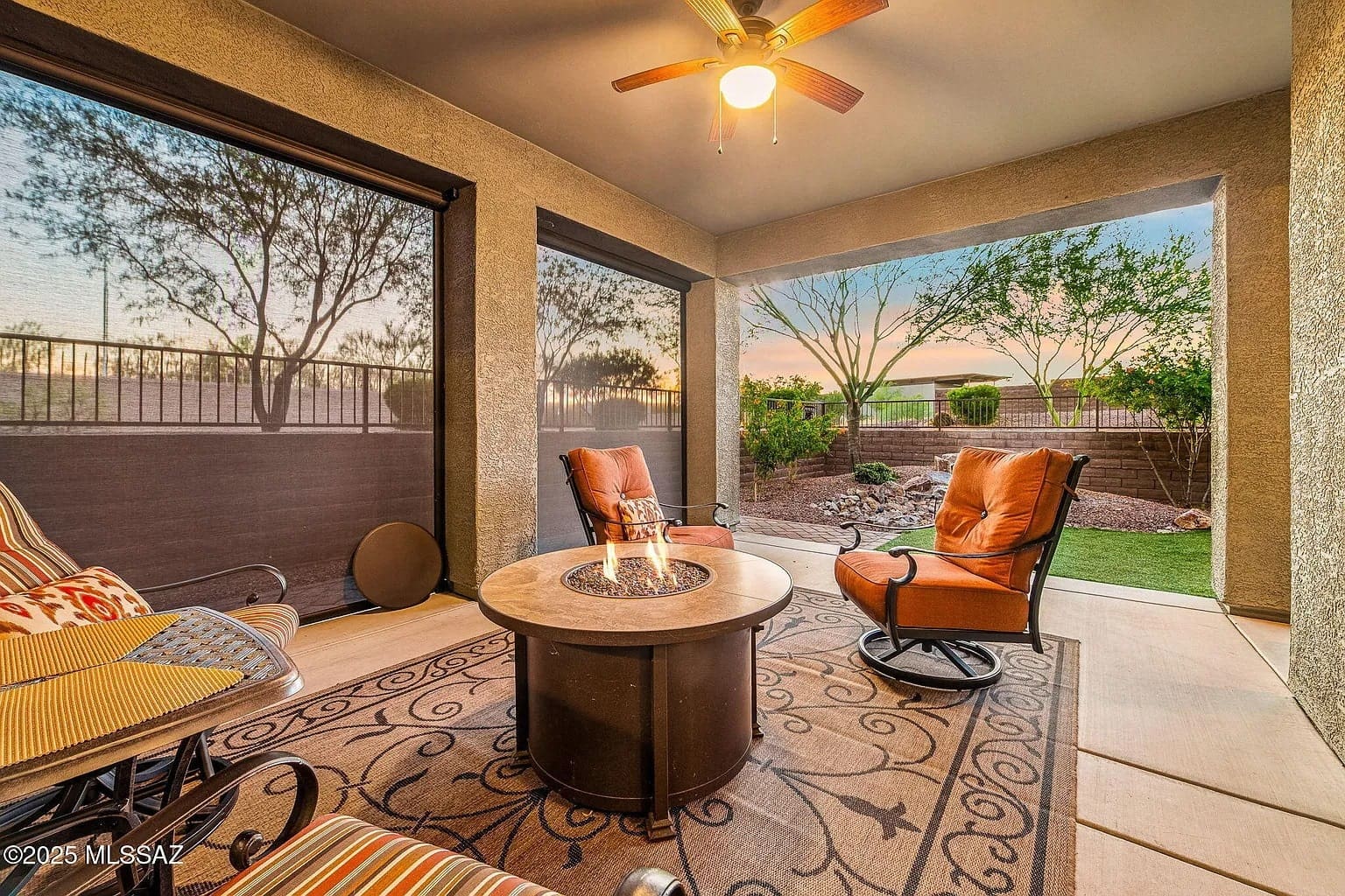 Covered patio with cushioned chairs, fire pit, ceiling fan, and view of a landscaped garden at sunset.