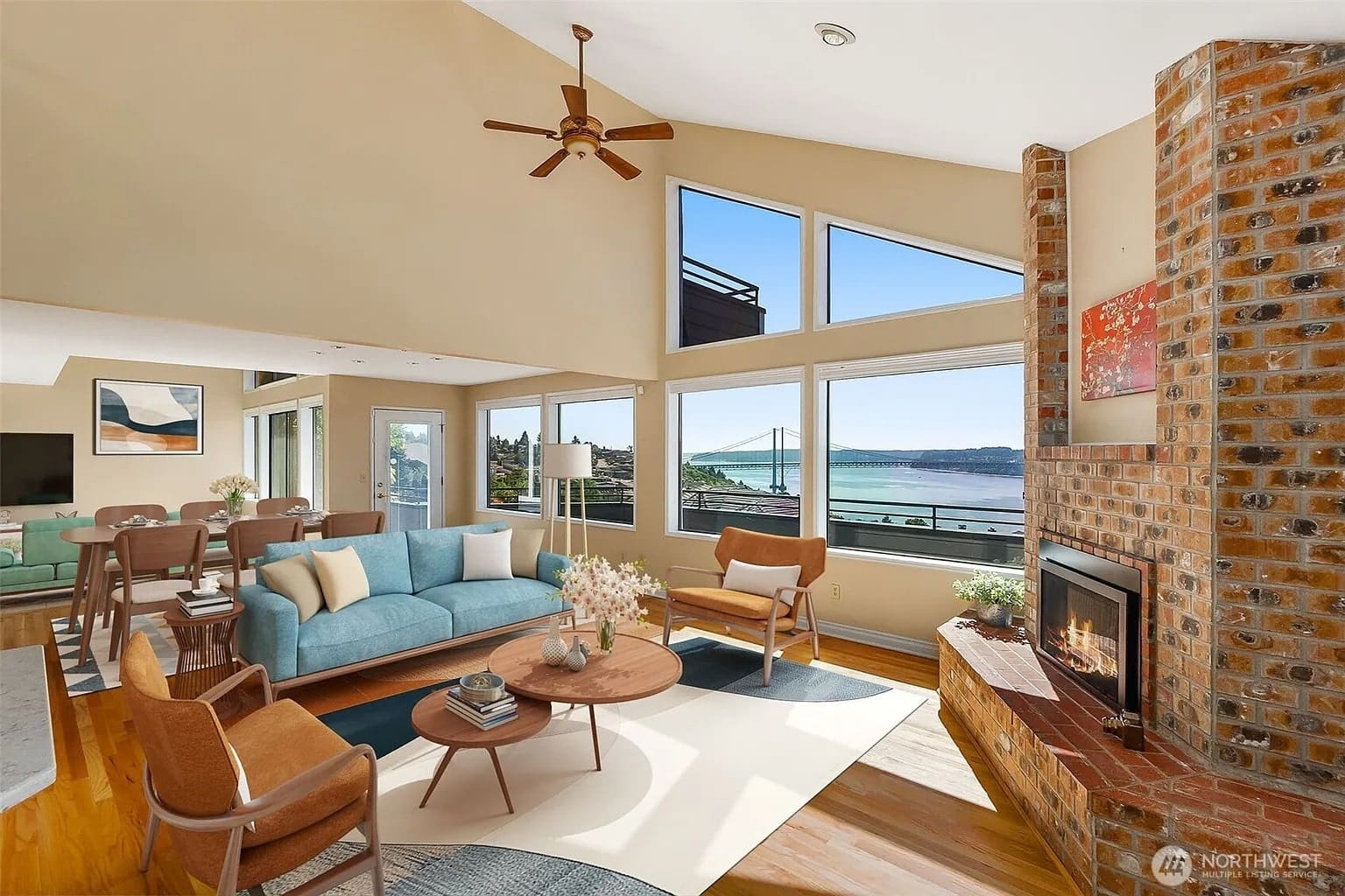 Spacious living room with large windows, blue sofa, armchairs, and a brick fireplace overlooking a bridge.