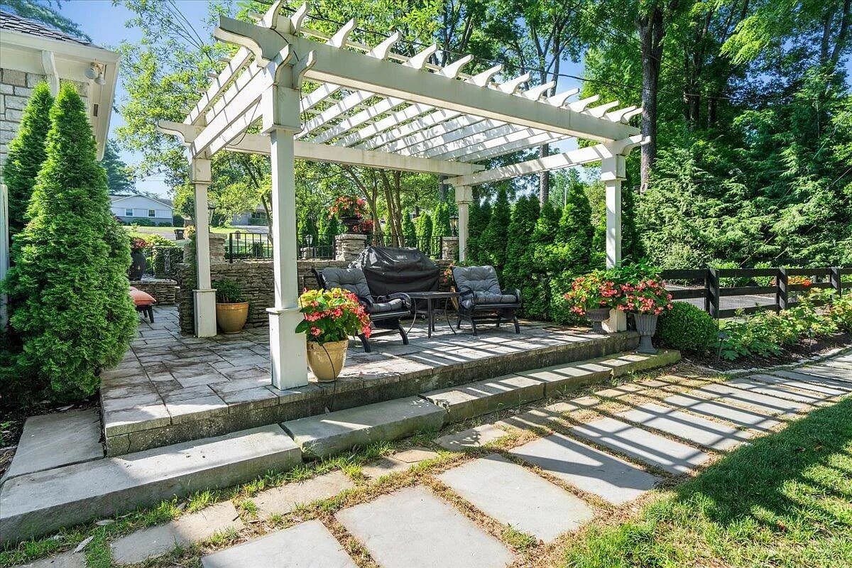 Outdoor patio with pergola, seating area, potted plants, and surrounding greenery.