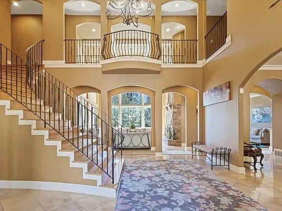 Spacious foyer with grand staircase, chandelier, and arched doorways leading to a bright living area.