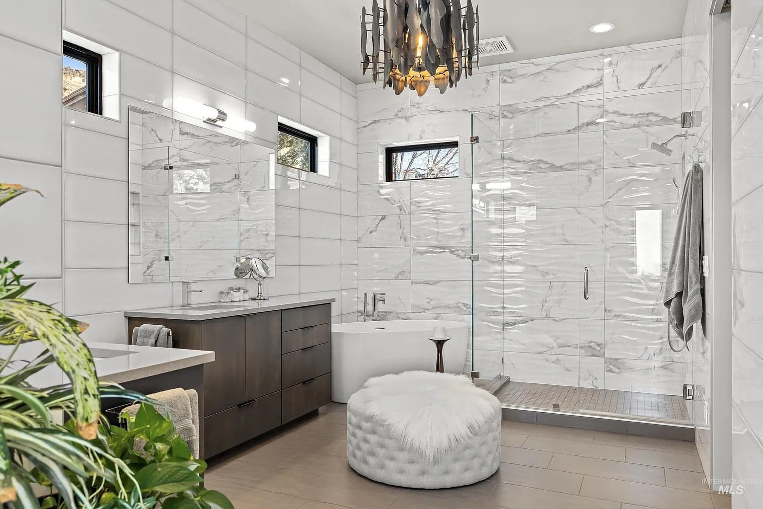 Modern bathroom with marble walls, glass shower, freestanding tub, vanity, and decorative chandelier.