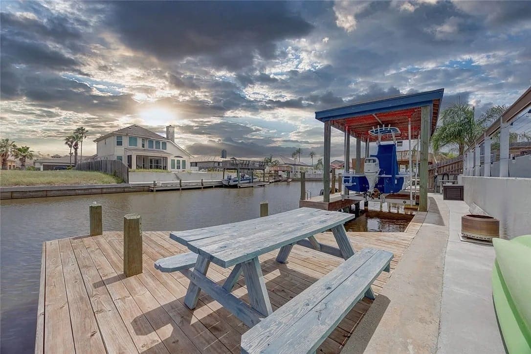 Canal-side wooden deck with a picnic table, boat lift, and houses under a cloudy sky.