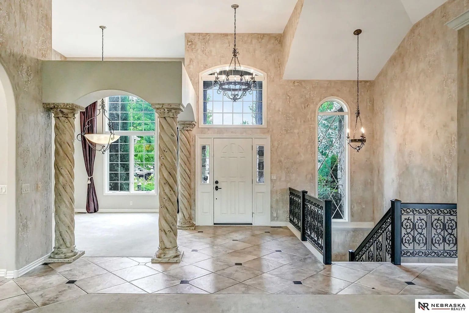Spacious foyer with high ceilings, chandelier, decorative columns, and large windows overlooking greenery.