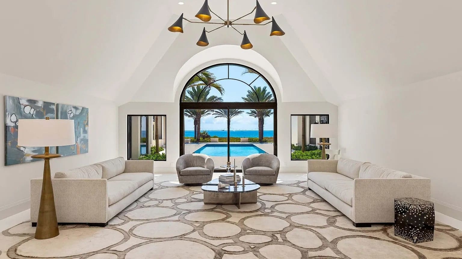Spacious living room with modern furniture, large arched window, pool, and ocean view.