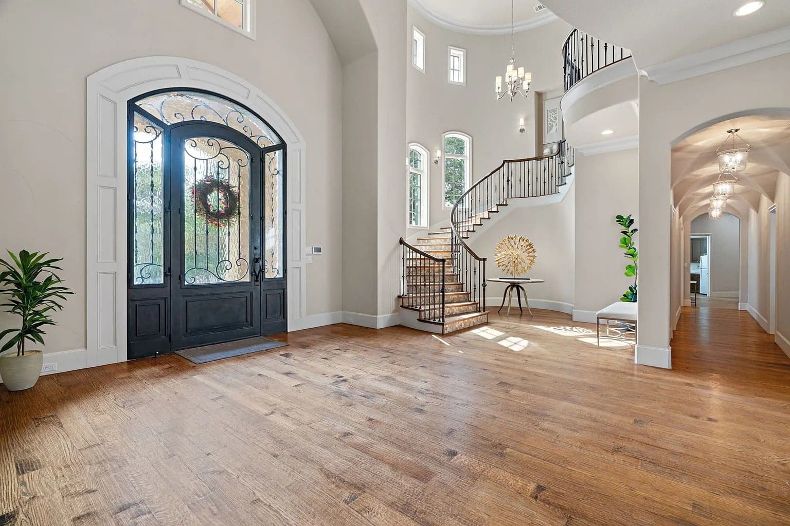 Spacious foyer with wooden floors, elegant staircase, decorative plants, and a large ornate front door.
