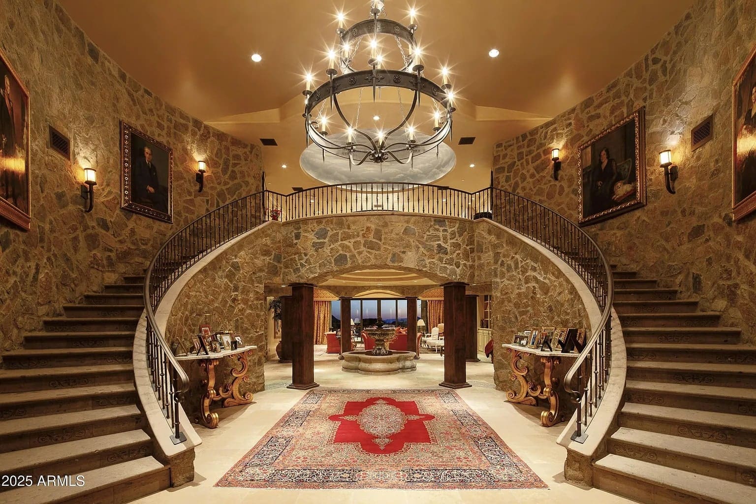 Grand foyer with dual staircases, stone walls, chandelier, and a central red-patterned rug.