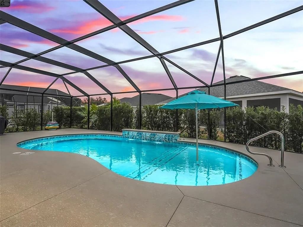 Enclosed swimming pool with a waterfall feature and umbrella, set against a colorful sunset sky.
