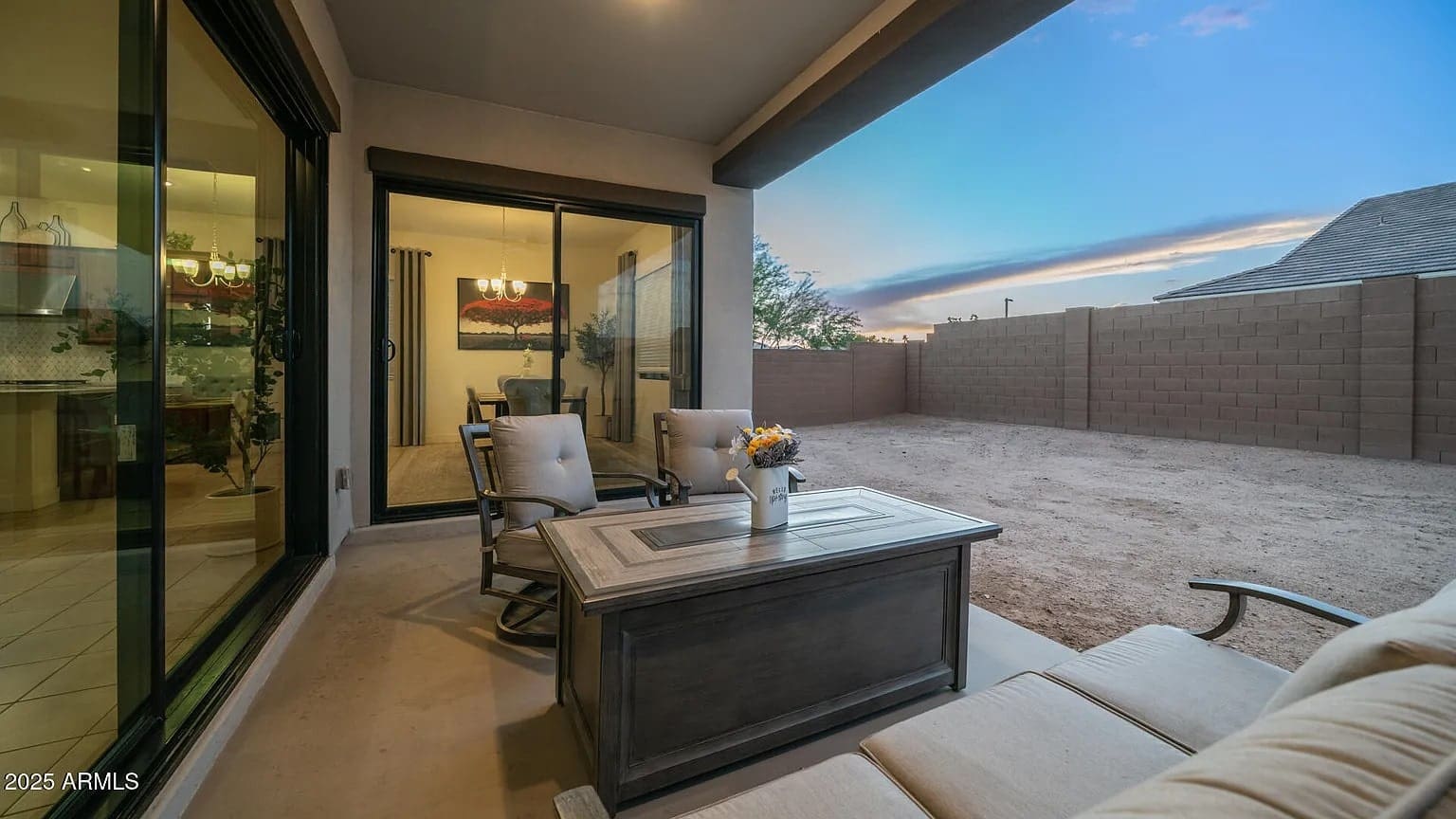 Covered patio with seating and table, overlooking a fenced dirt yard at sunset.