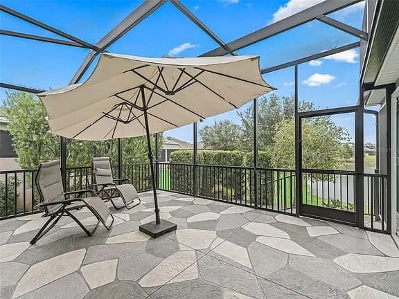 Screened patio with two lounge chairs, large umbrella, and a view of greenery outside.