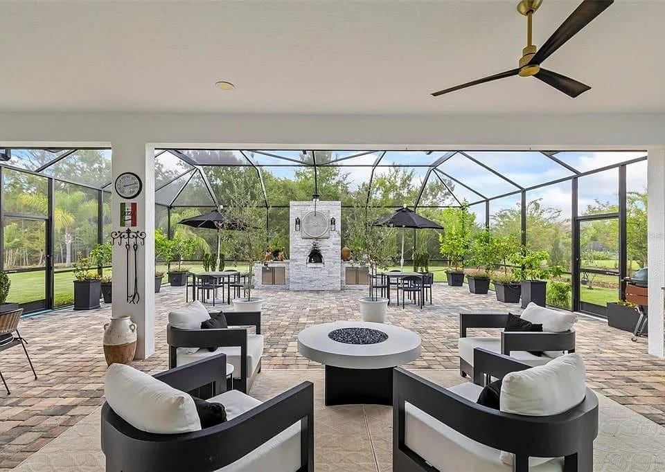 Spacious outdoor patio with modern furniture, fireplace, ceiling fan, and large windows overlooking greenery.