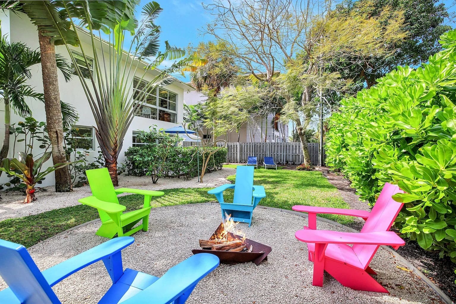 Colorful chairs around a fire pit in a lush backyard with trees and a house.