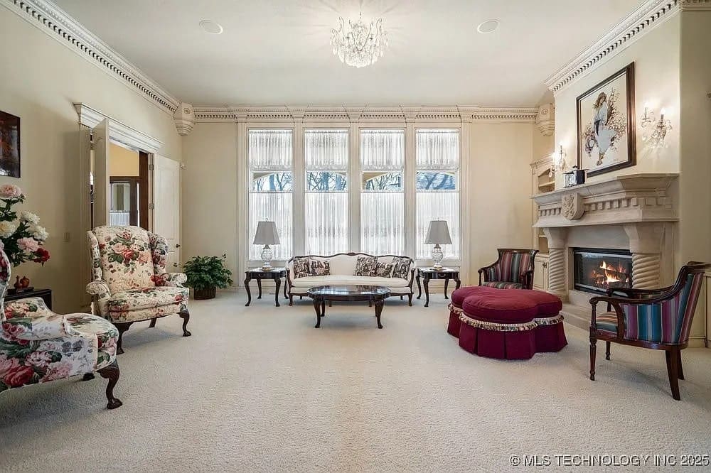 Elegant living room with floral armchairs, ornate fireplace, chandelier, and large windows with sheer curtains.