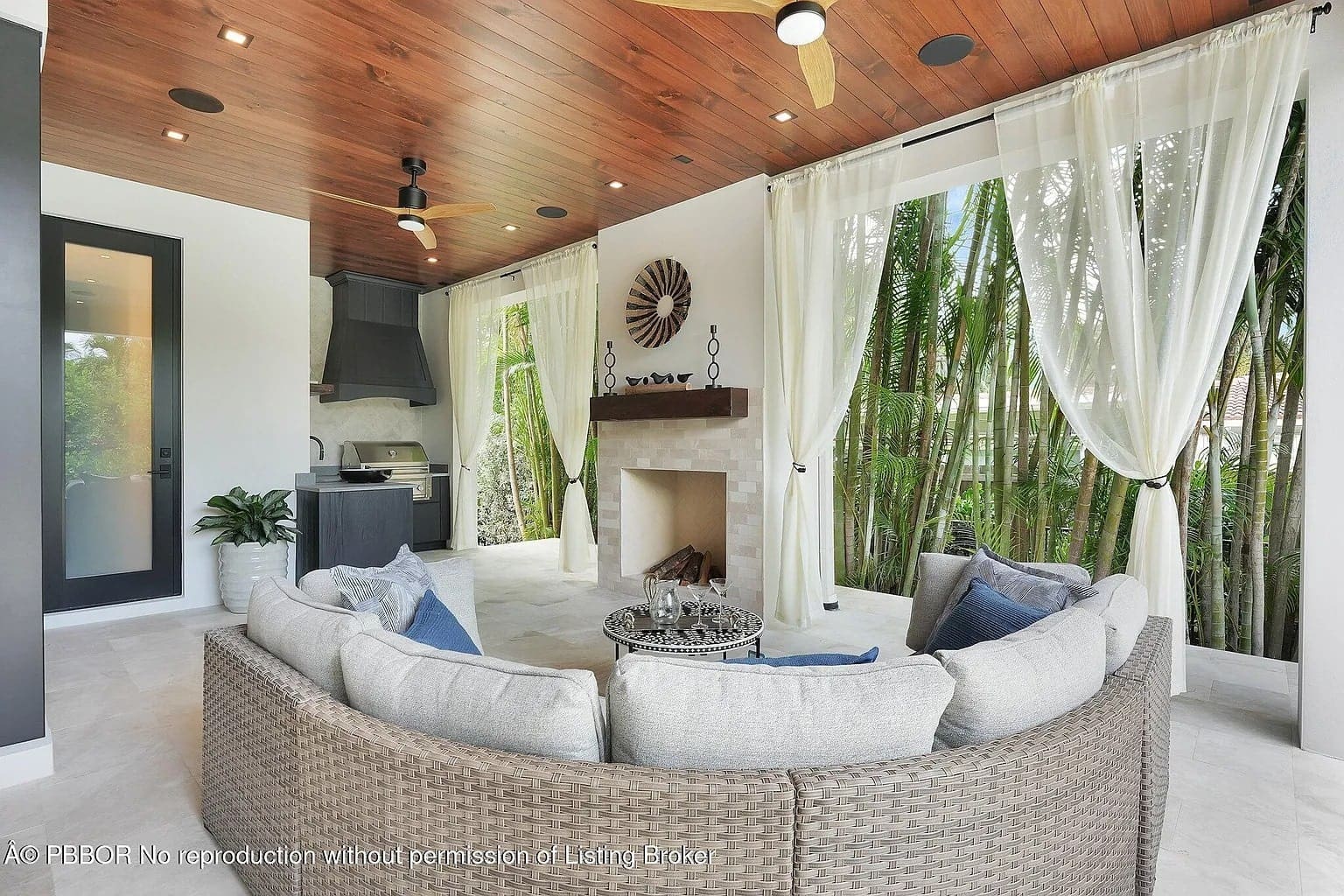 Outdoor patio with a circular sofa, fireplace, and curtains, surrounded by tropical plants.