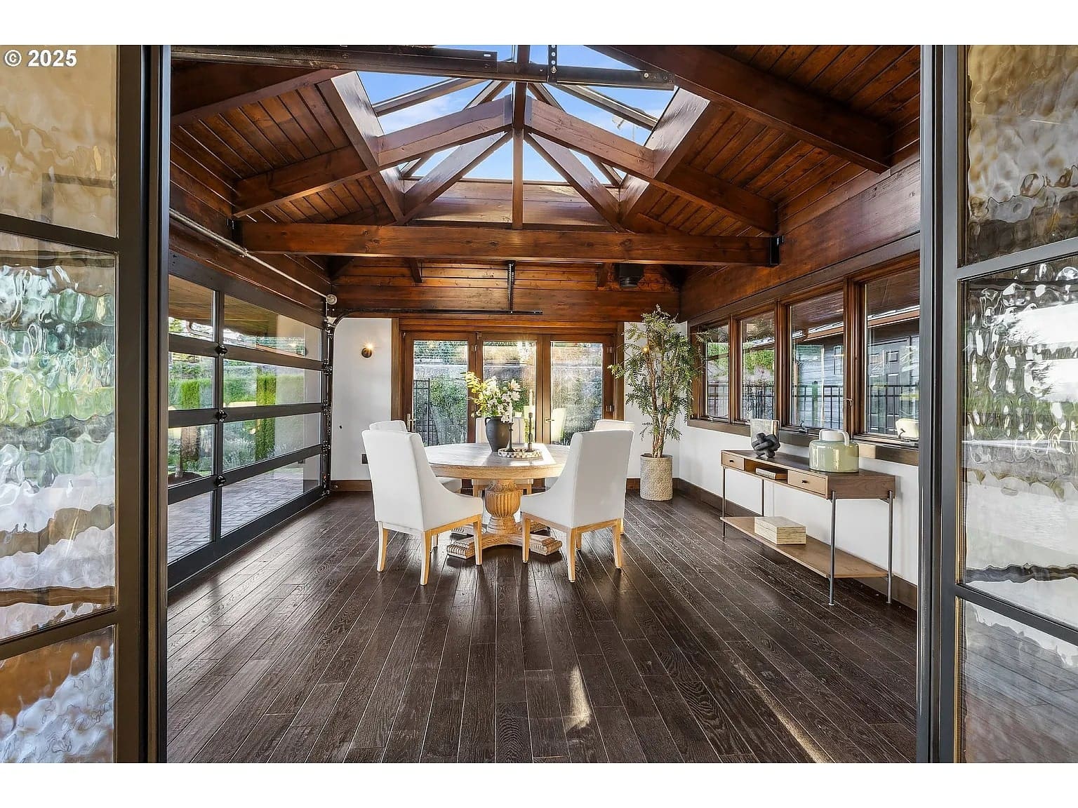 Wooden dining room with skylight, round table, white chairs, and large windows offering natural light.