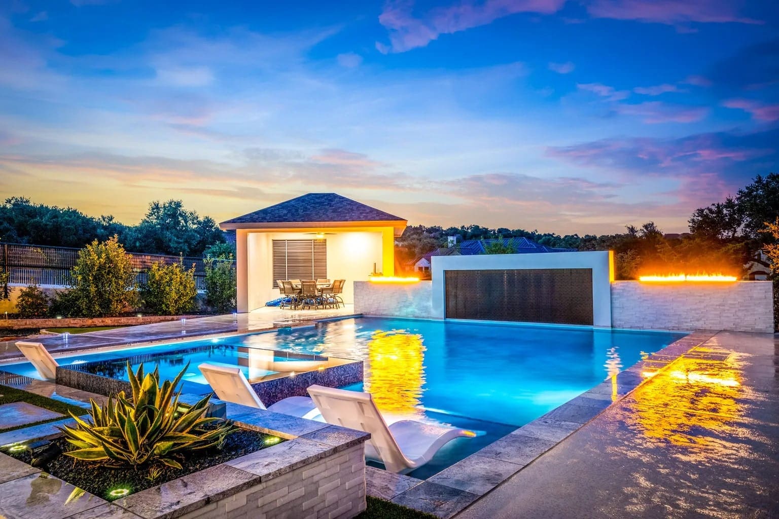 Modern backyard with illuminated pool, waterfall feature, lounge chairs, and a small pavilion at sunset.

