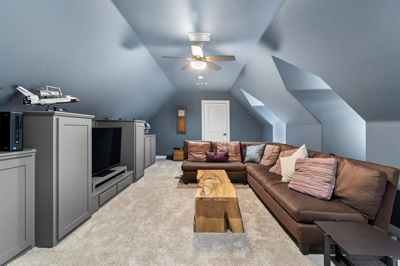 Attic living room with brown sectional sofa, wooden coffee table, and large TV under sloped ceiling.
