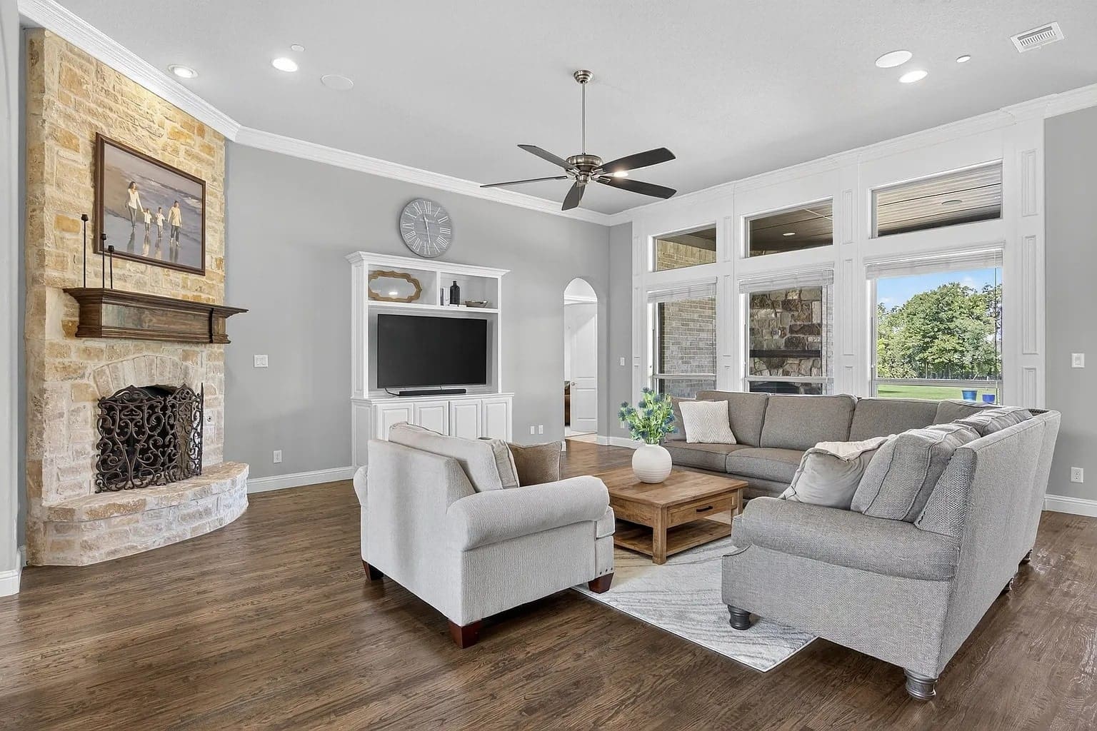 Modern living room with gray sofas, stone fireplace, TV, ceiling fan, and large windows.

