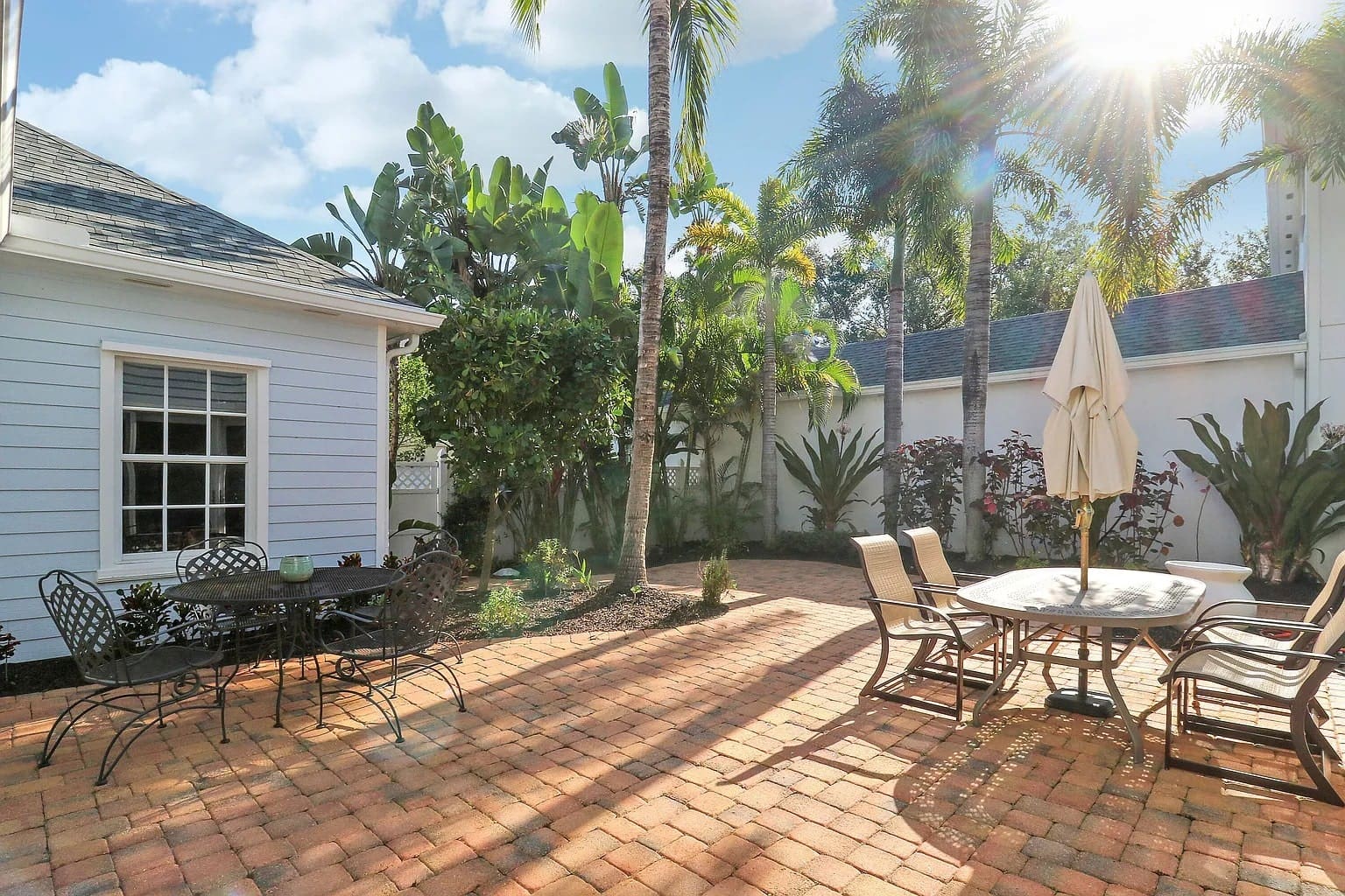 Sunny backyard patio with tables, chairs, palm trees, and a white house.
