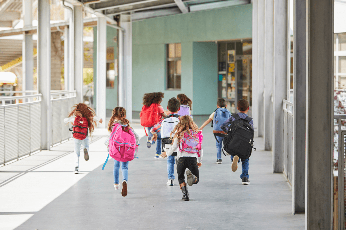 A group of young children with backpacks joyfully run down a school hallway, heading towards a classroom.