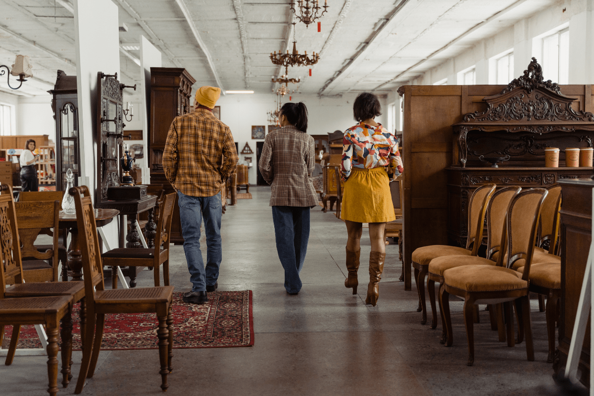 Three people walk through a spacious antique store with vintage furniture, including ornate wooden chairs and tables, displayed along the aisle.
