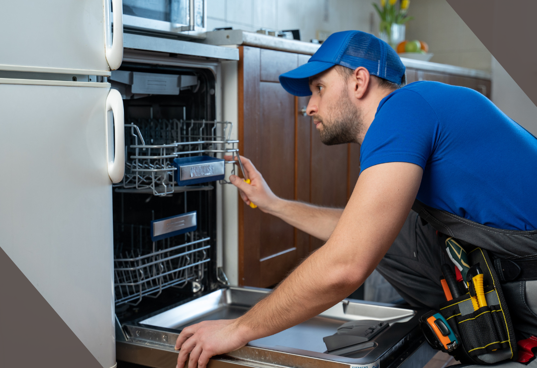 Man in blue cap and shirt repairing a dishwasher in a kitchen.