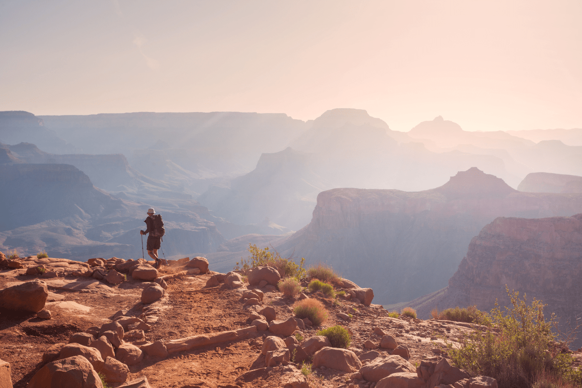 A lone hiker stands on a rocky path overlooking the expansive, sunlit Grand Canyon, with layers of hazy cliffs and valleys stretching into the distance.