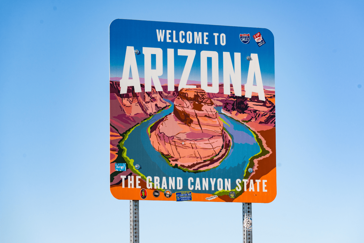 A colorful sign welcomes visitors to Arizona, featuring a stylized illustration of the Grand Canyon with the text "The Grand Canyon State" against a clear blue sky.