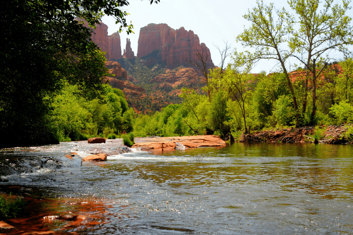 A tranquil river flows through lush greenery with red rock formations towering in the background under a clear sky.