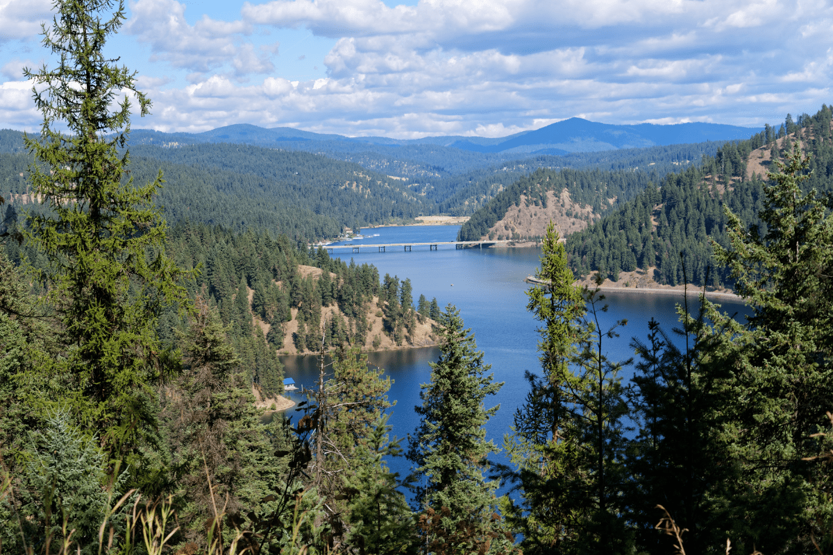 A scenic view of a serene lake surrounded by dense forests and rolling hills under a partly cloudy sky, with a bridge visible in the distance.