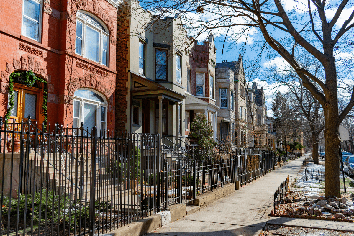 A row of colorful, historic brick townhouses with decorative iron fences lines a tree-lined sidewalk under a clear blue sky.