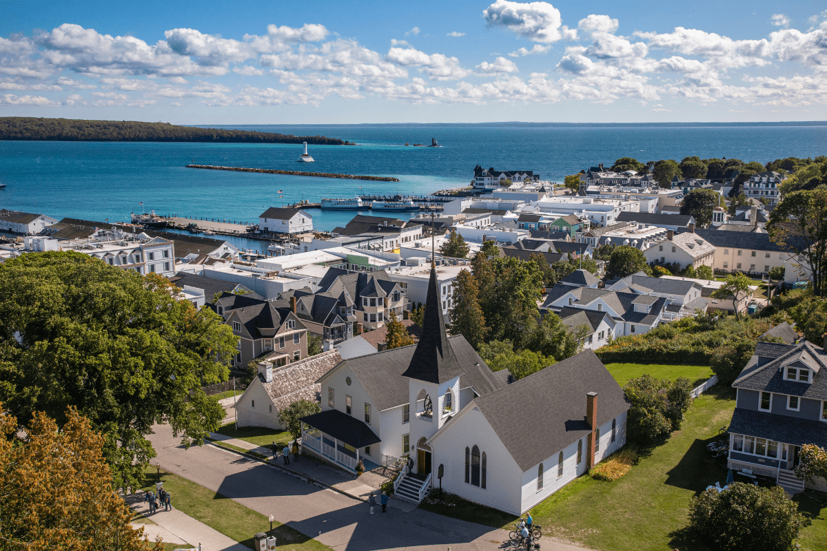 Aerial view of a coastal town with a prominent church, surrounded by houses, overlooking a vibrant blue sea with a lighthouse and breakwater under a partly cloudy sky.