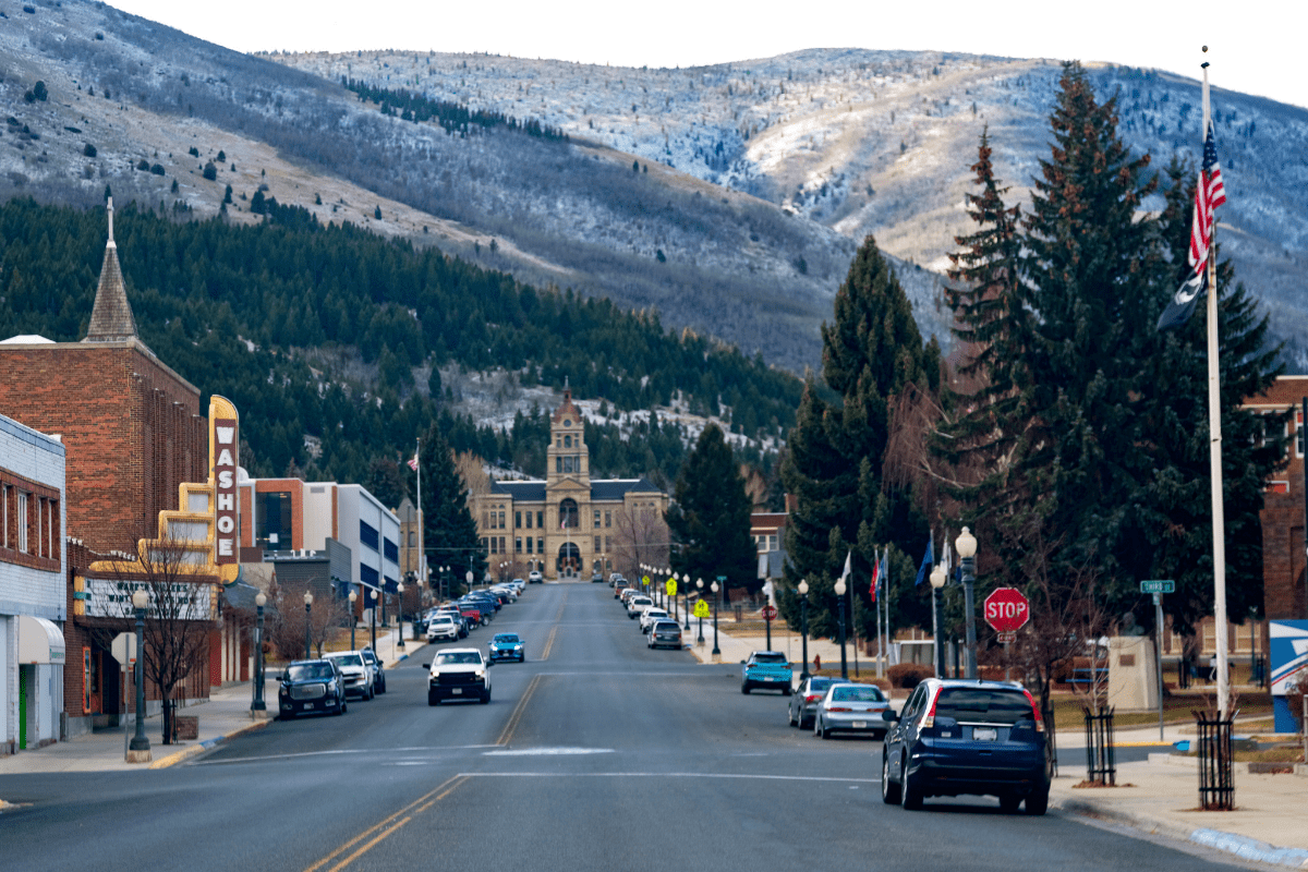 A small town street lined with parked cars and buildings leads to a distant courthouse, set against a backdrop of snow-dusted mountains and evergreen trees.