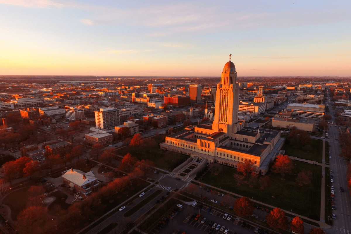 Aerial view of a cityscape at sunset, featuring a prominent tall building with a dome, surrounded by smaller buildings and streets, bathed in warm, golden light.