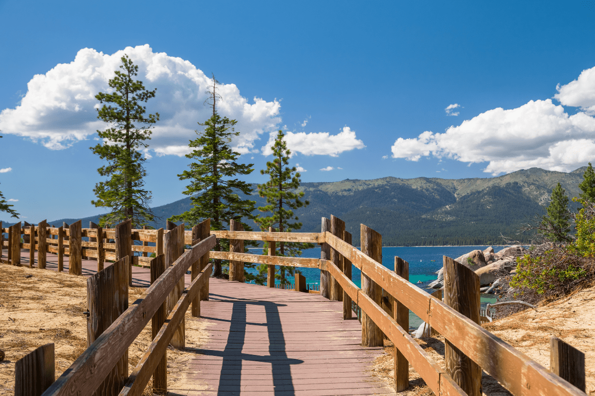 A wooden boardwalk with railings leads through a sandy area with pine trees, overlooking a blue lake and distant mountains under a clear sky with scattered clouds.