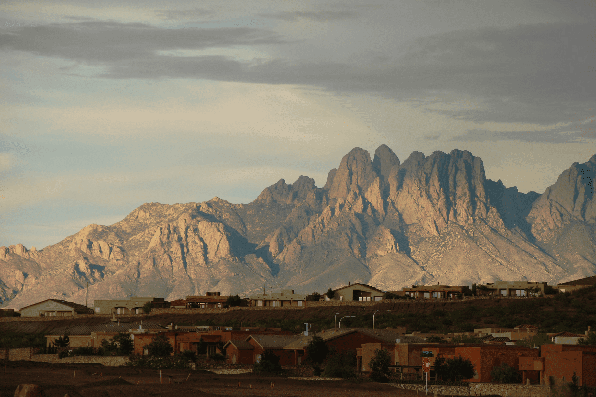 A scenic view of rugged mountains under a cloudy sky, with a residential area of adobe-style houses in the foreground.