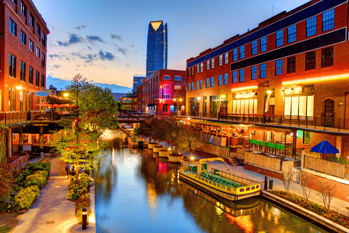 A vibrant canal scene at sunset features colorful buildings, a boat docked by the water, and a distant modern skyscraper under a partly cloudy sky.