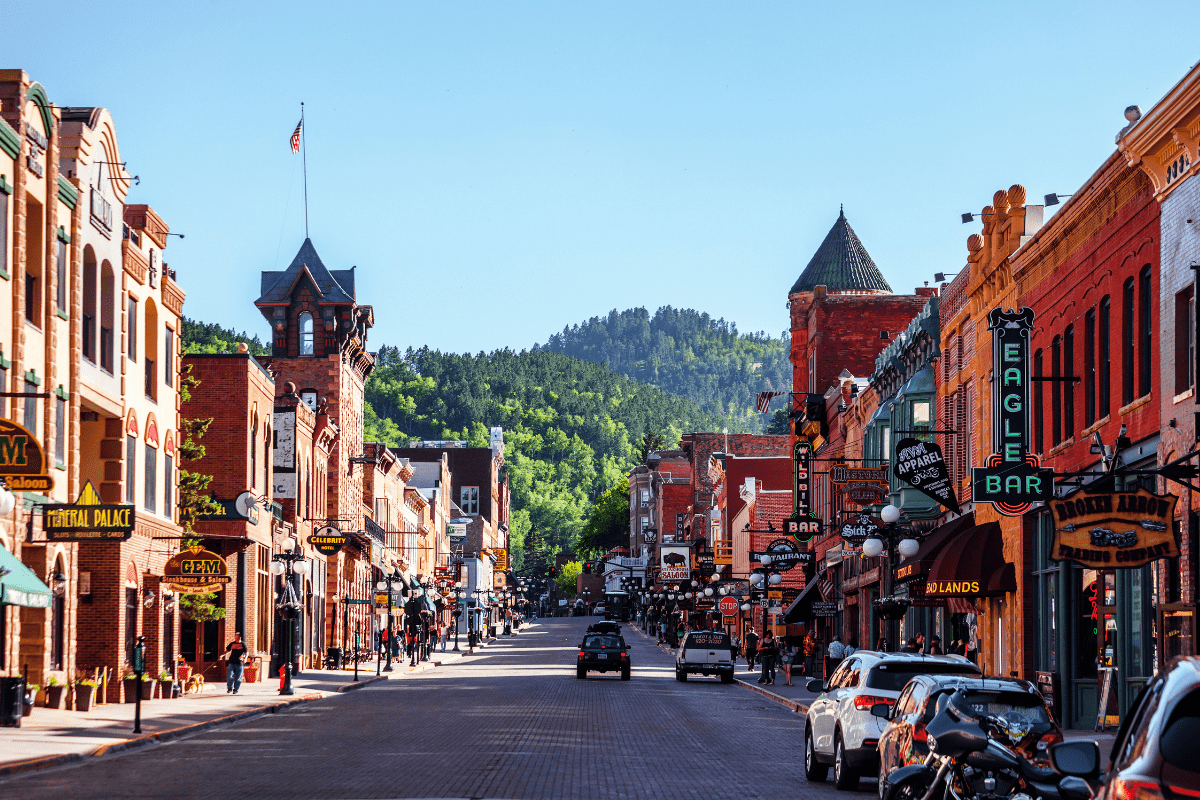 A vibrant street scene in a historic town features colorful brick buildings, vintage signs, parked cars, and lush green hills in the background under a clear blue sky.